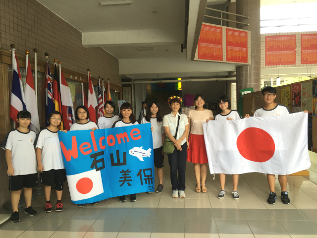 日本語交流をお手伝いしてくれる生徒たちと記念の集合写真。「ウェルカム石山美保さん」と書かれた横断幕と国旗を持っている