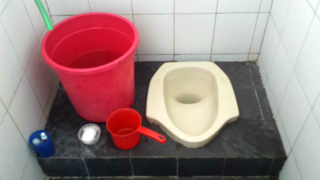 和式便所に似ている便器に、水が入った大きな赤い桶と手桶、固形石鹸が置いてある写真