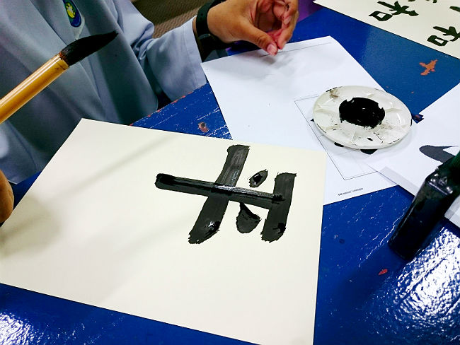 日本語パートナーズ マレーシア ジョホール 書道本番は半紙に「平和」と漢字を書いている写真 