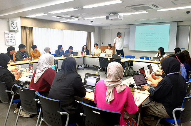 The classroom is set up with desks arranged in a square. The presenter stands in front of the screen, and the participants pay close attention.