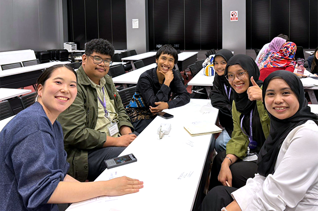 In a classroom with neatly arranged long white desks, participants are sitting around one desk and smiling at the camera.