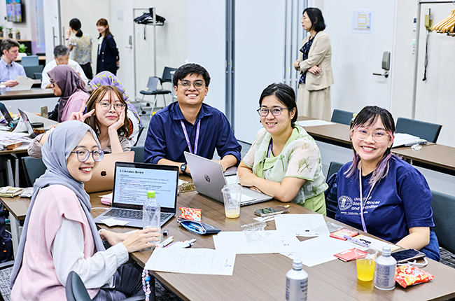 Five participants sit around two long tables in a corner of the classroom, smiling at us.