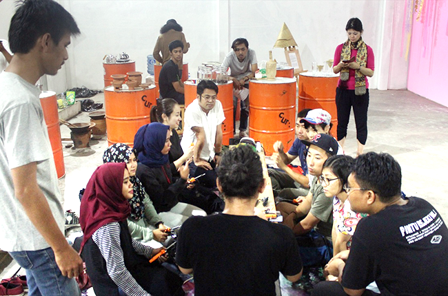 白い壁の部屋で、板をテーブル代わりにして10人ほどの人々が座っておしゃべりしている様子が映っており、背後の壁際にドラム缶が置かれている。