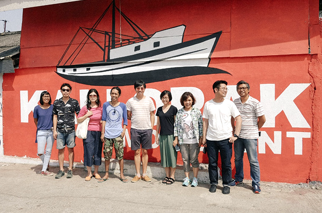船のイラストが描かれたオレンジ色の壁の前に9人が並んで記念撮影している様子