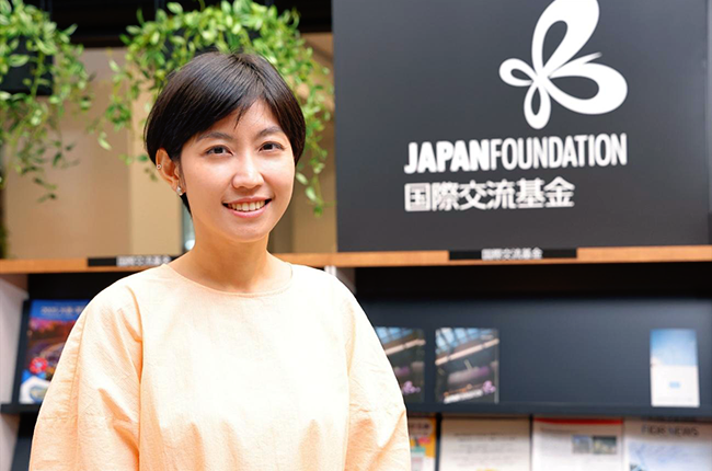 国際交流基金1階の図書館にあるJFのロゴが入った看板の前で、こちらを見ているレ・トゥアン・ウイエンさん