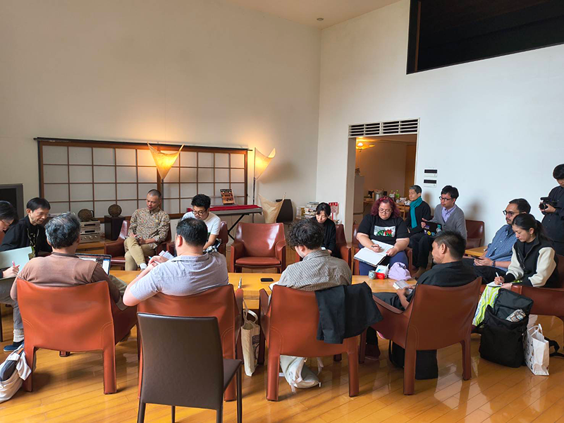 Fifteen people are sitting in chairs around a table.