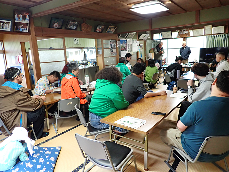A man is standing in a Japanese-style room, explaining something to 15 people.