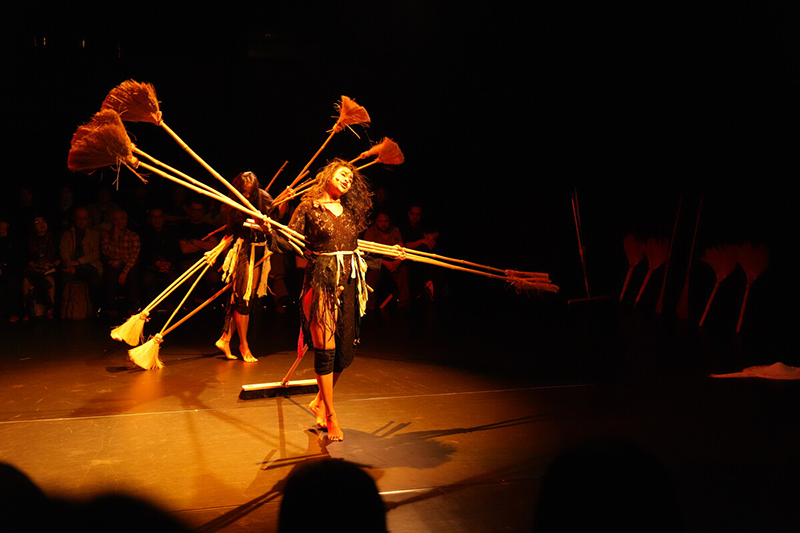 There are two women on stage, each holding a broom.