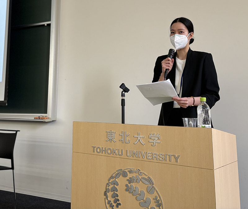 口にマスクをしたスーツ姿の女性が東北大学の壇上で講演している。