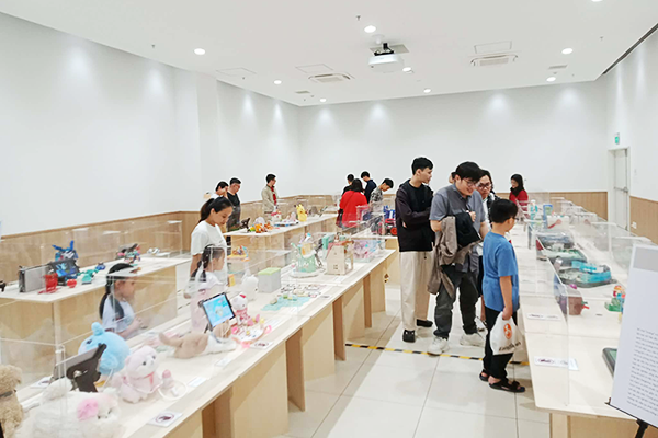 ぬいぐるみやパーティーゲーム、フィギュアなどのおもちゃがガラスケース内に展示されている。来場者が15名ほど写っている。