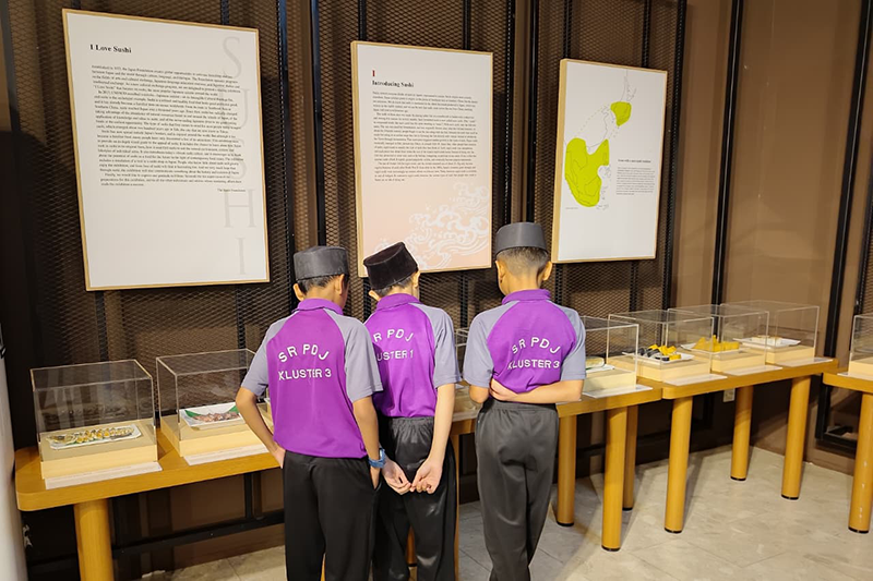 寿司についての展示を鑑賞している男の子3人の背中が写っている。