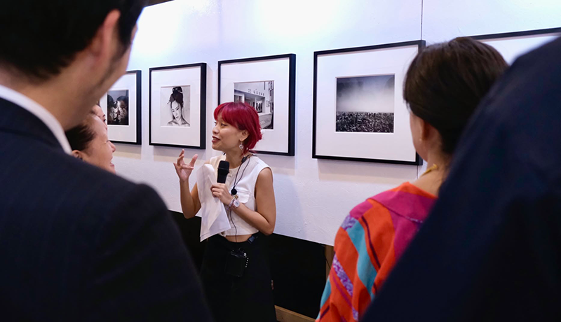 壁に展示されているモノクローム写真の前で1人の女性がマイクを持って説明している姿を見ている鑑賞者の後ろ姿。